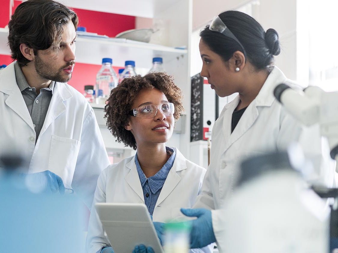 Multi-ethnic scientists discussing over digital tablet. Male and female researchers working together in laboratory. They are in uniforms.